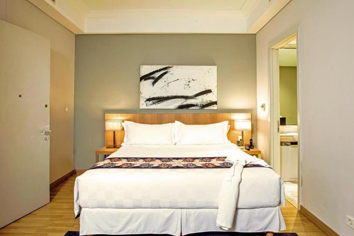 A hotel room with a bed, two bedside lamps, a large abstract painting on the wall, and a wooden floor.