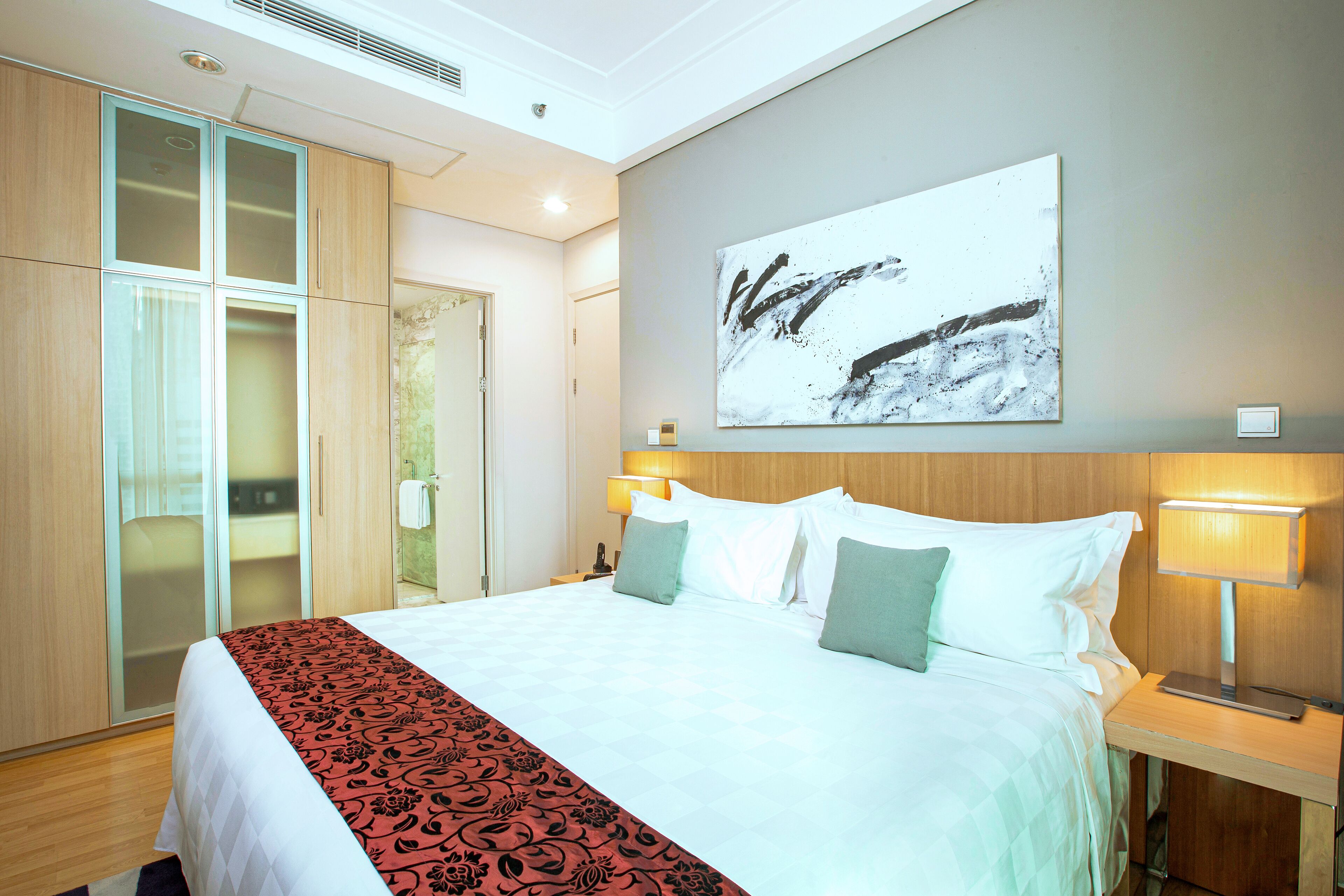 A hotel room with a large bed, a wardrobe, a bedside table with a lamp, and a wall-mounted artwork.