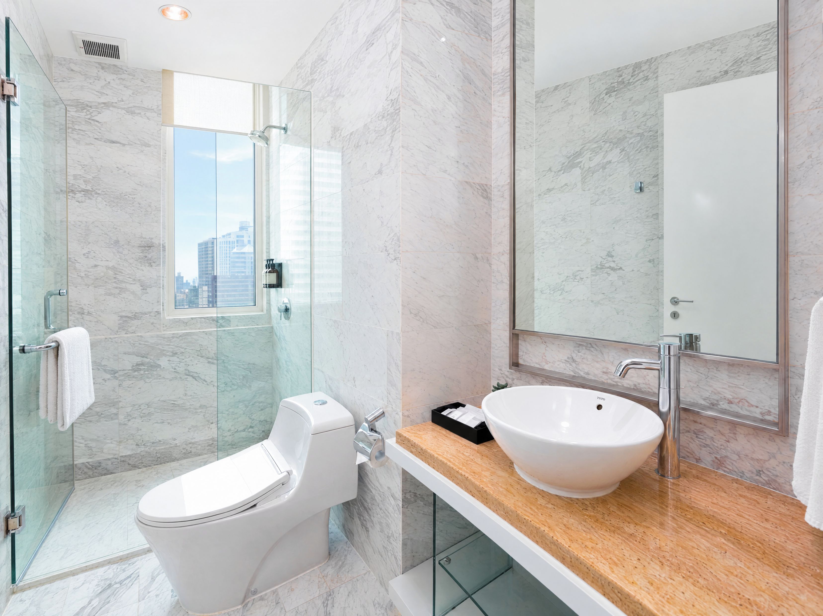 A modern bathroom with a glass-enclosed shower, a white toilet, a large sink with a mirror, and a view of the city through the shower door.