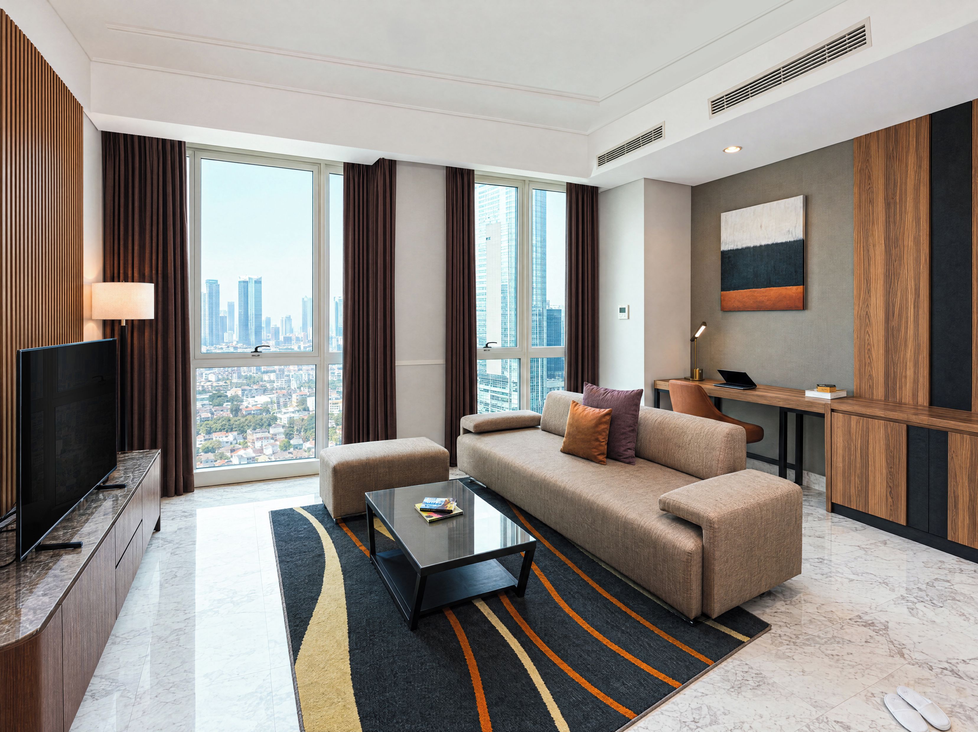 A modern hotel room with a large window, a flat-screen TV, a sofa, a coffee table, and a desk with a lamp.
