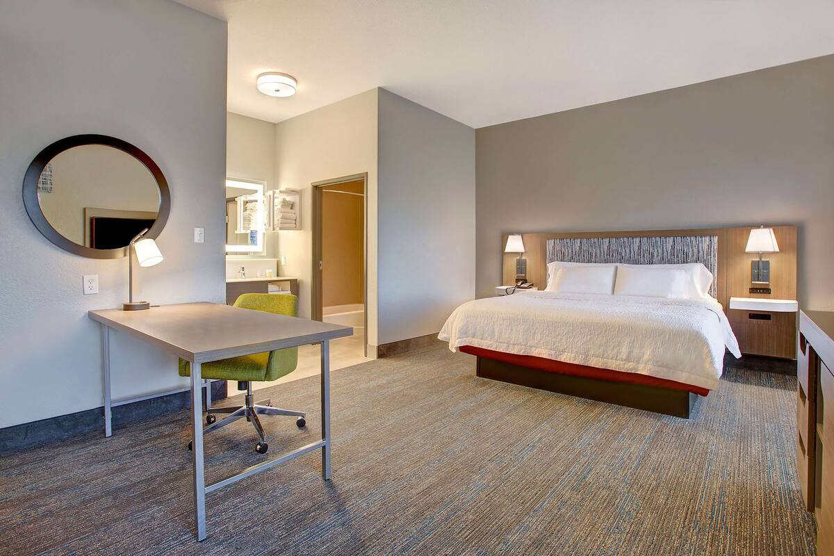 A hotel room with a bed, a desk, a chair, a mirror, and a bathroom visible through an open door.