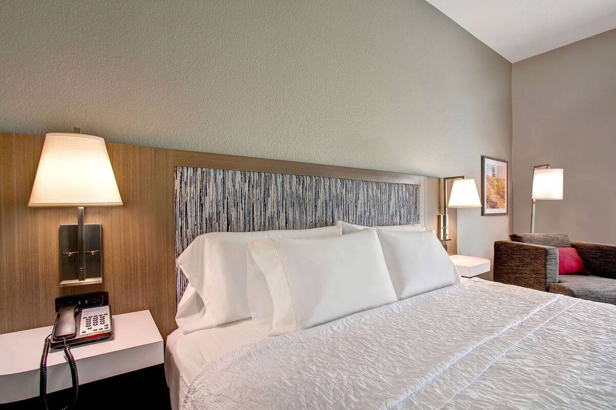 A hotel room with a large bed, a bedside table with a phone, a lamp, and a couch with a red pillow.