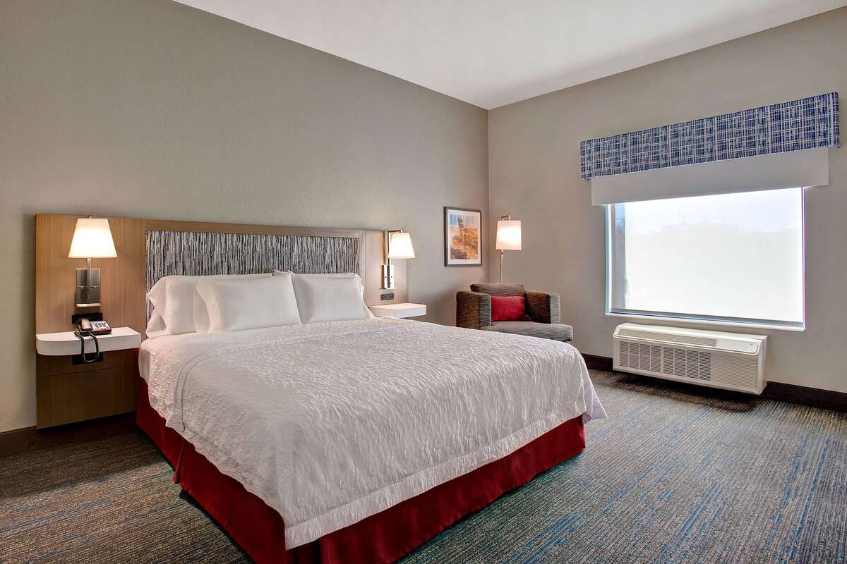 A hotel room with a large bed, two bedside tables with lamps, a window with blinds, and a wall-mounted air conditioner.