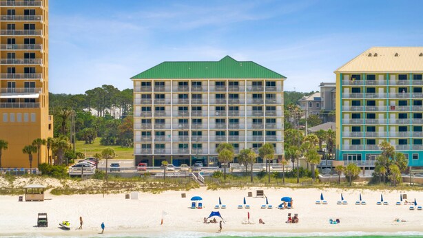 Een hotel aan het strand met meerdere verdiepingen, een groen dak en een strandgedeelte met parasols en ligstoelen.