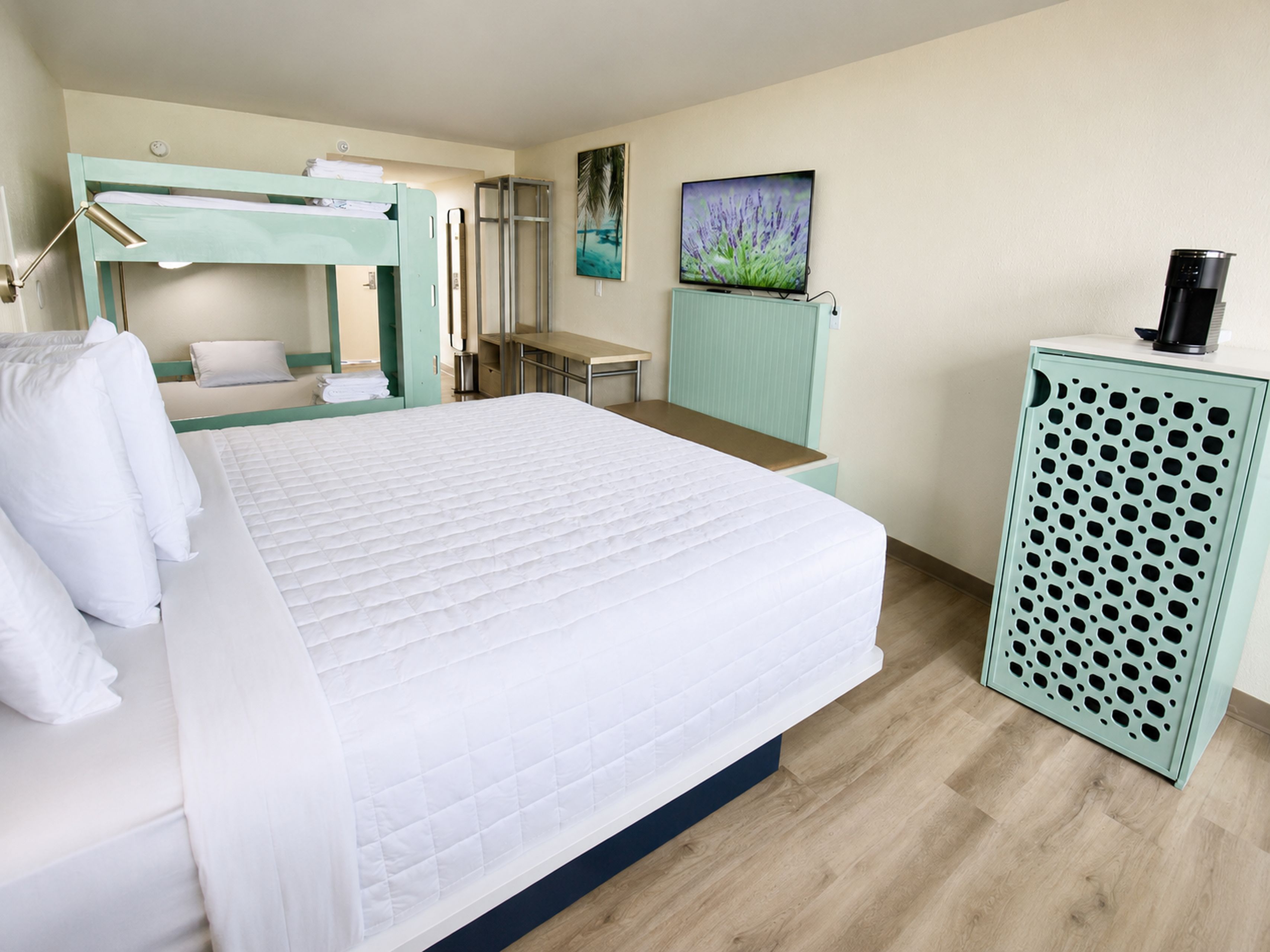 A hotel room with a bed, a desk, a bunk bed, a mirror, a painting, and a coffee machine.
