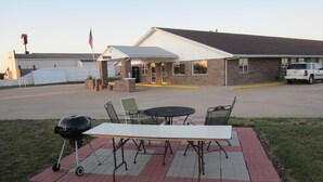 Un patio al aire libre con una parrilla, una mesa y sillas, frente a un edificio de fachada de ladrillo y toldo blanco.