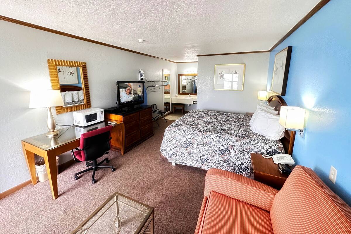 A hotel room with a bed, a desk with a computer, a chair, a microwave, a telephone, and a small table.