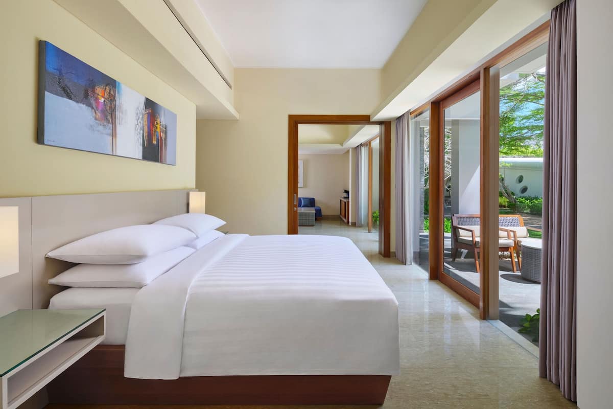 Quarto de hotel com cama grande, mesa de cabeceira, um quadro na parede e vista para a área externa por uma porta de vidro deslizante.