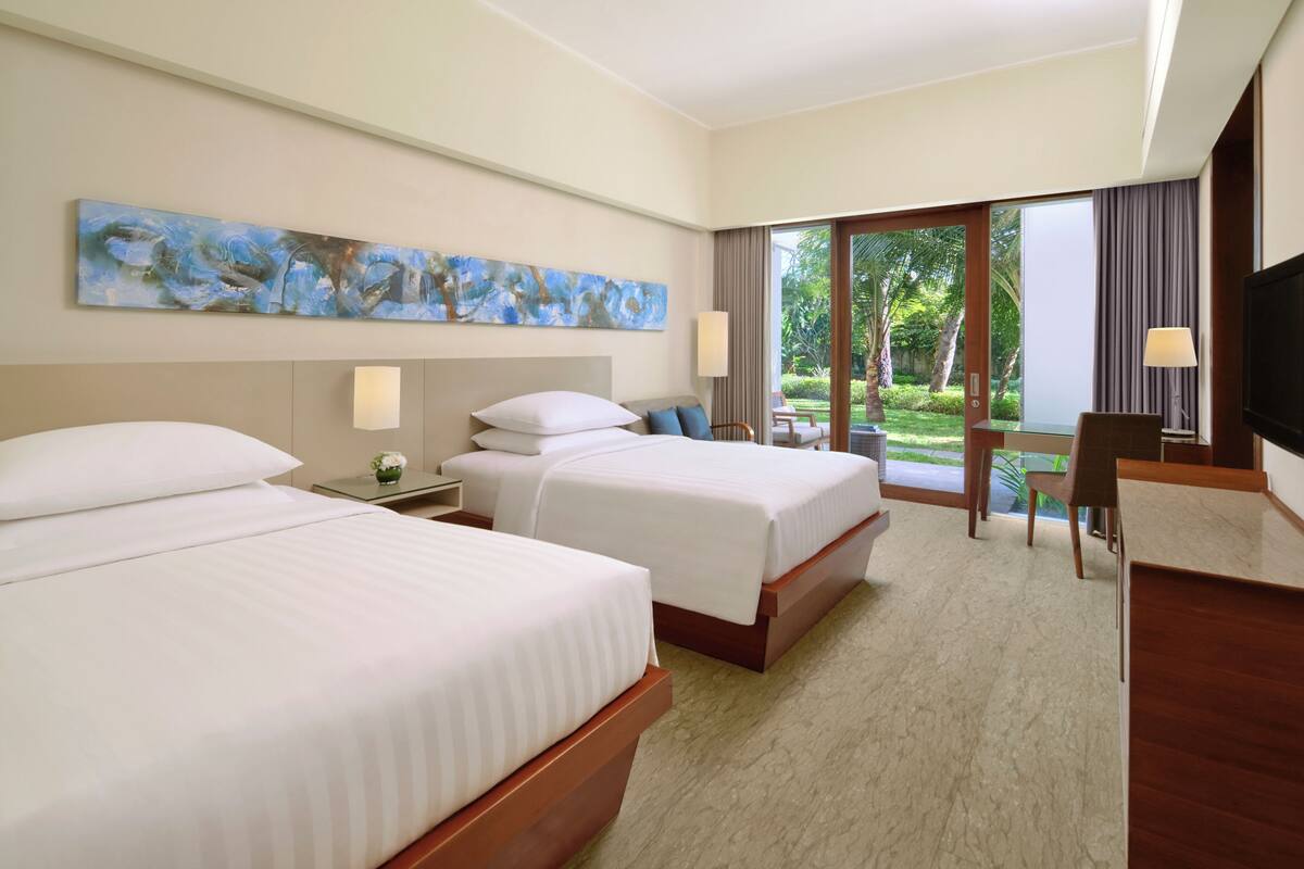 Quarto de hotel com duas camas, televisão de tela plana, escrivaninha e uma janela grande com vista para área verde.