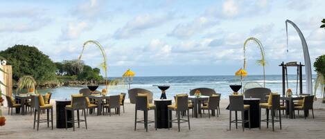 A beachside restaurant with outdoor seating, a view of the ocean, and a curved architectural feature.