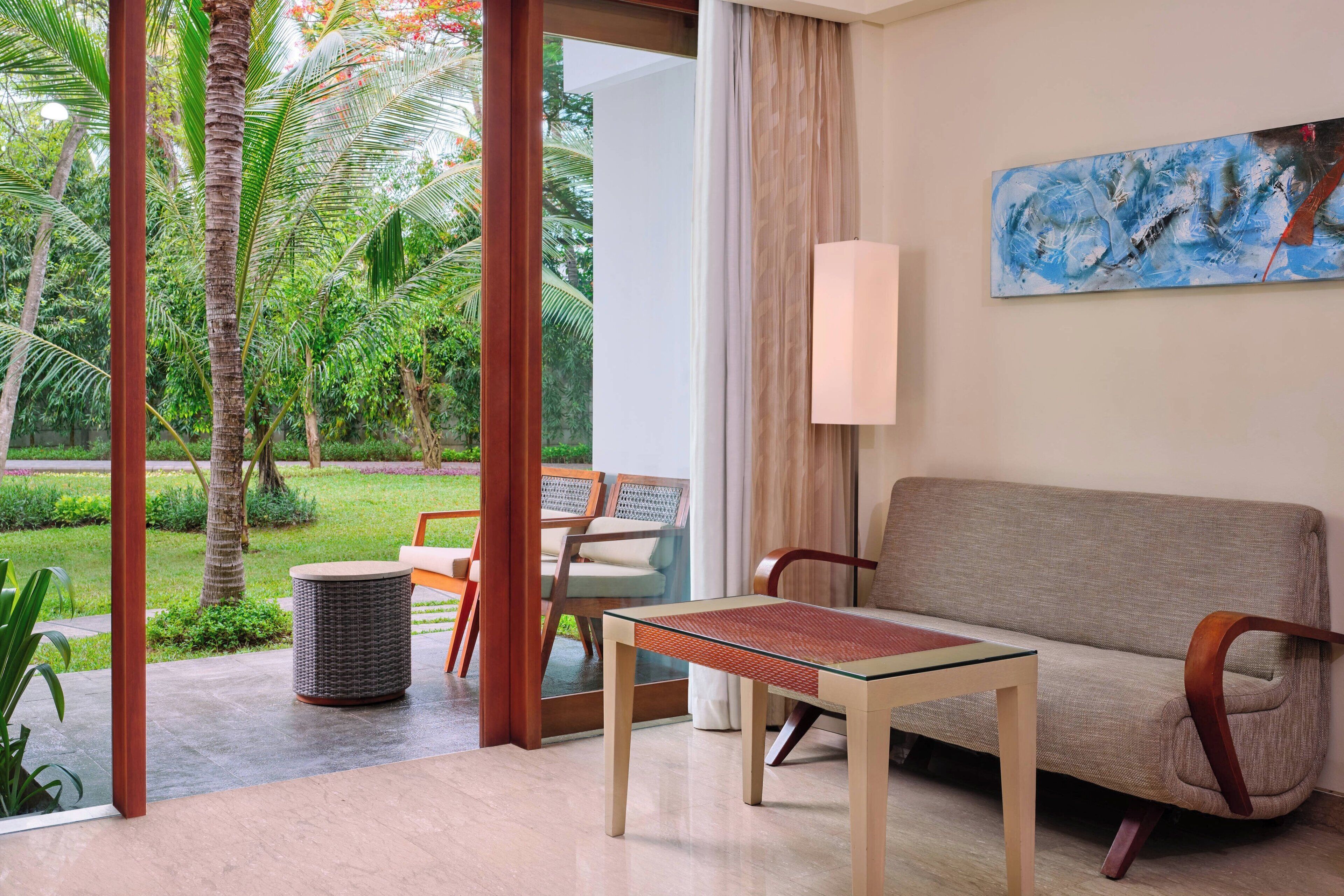 A modern hotel room with a sofa, a glass-top table, a lamp, and a view of a lush garden.