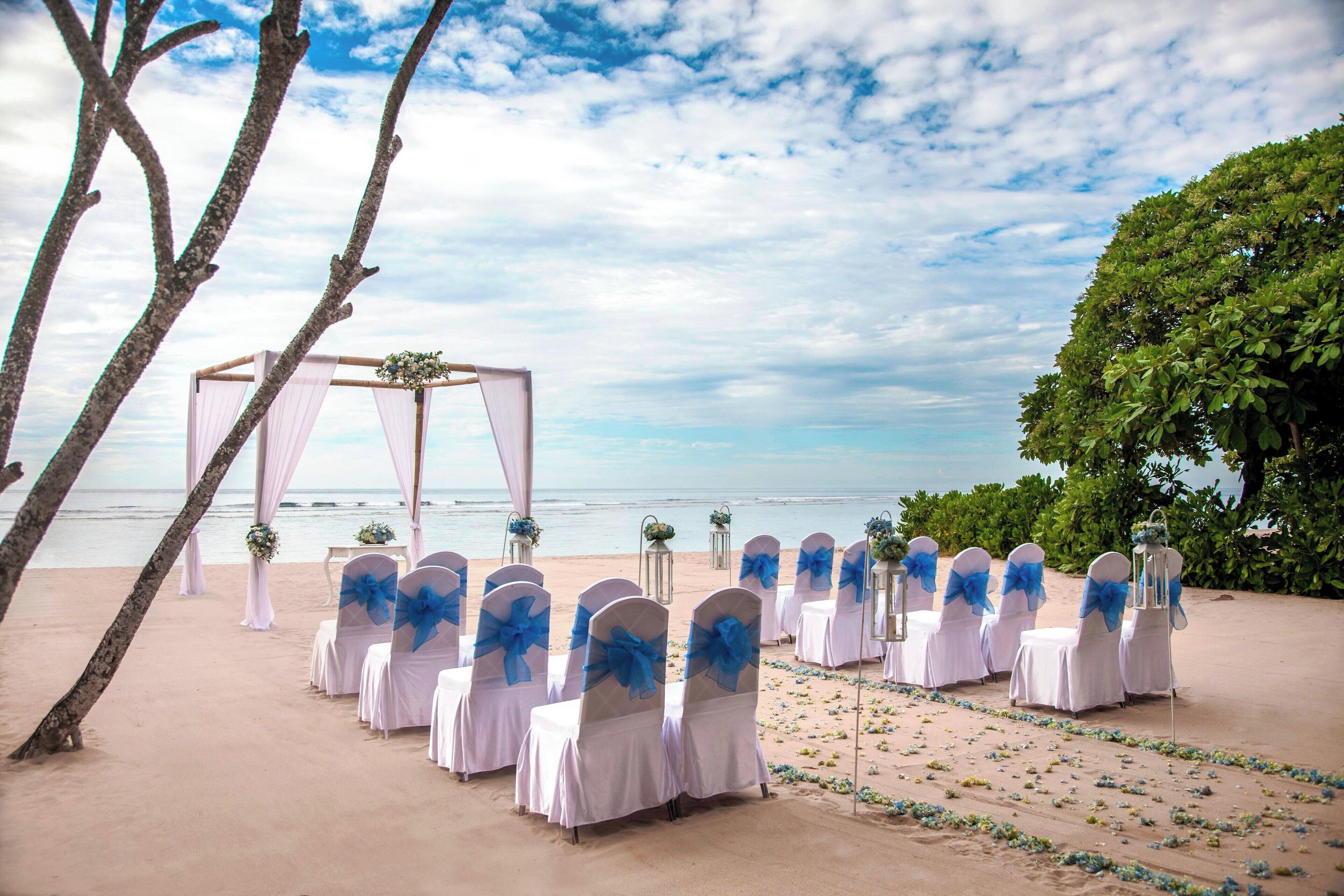 A beach wedding setup with white chairs and blue sashes, a floral arch, and tables with white tablecloths.