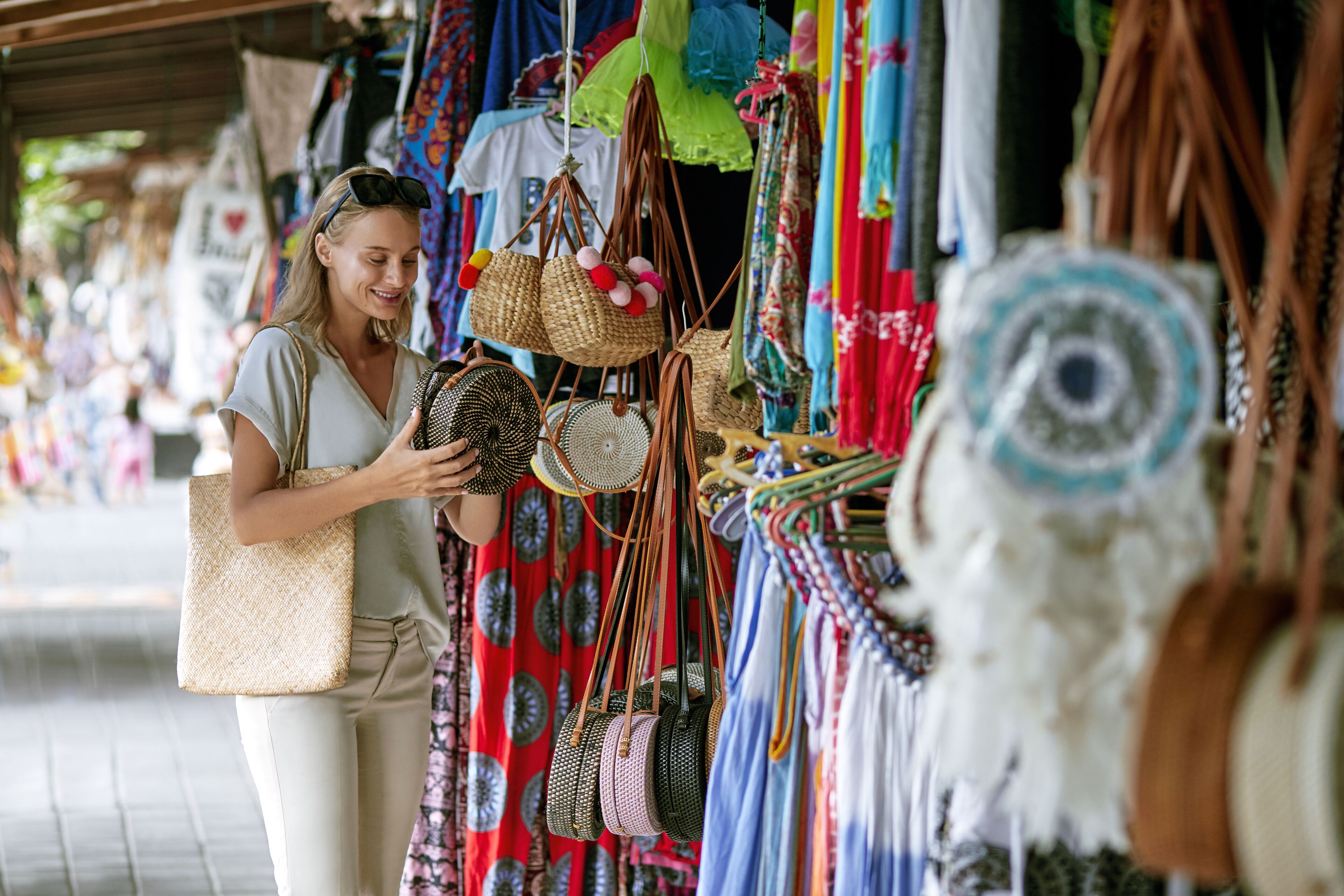 A person examining woven bags at a market stall with colorful fabrics and accessories in the background.
