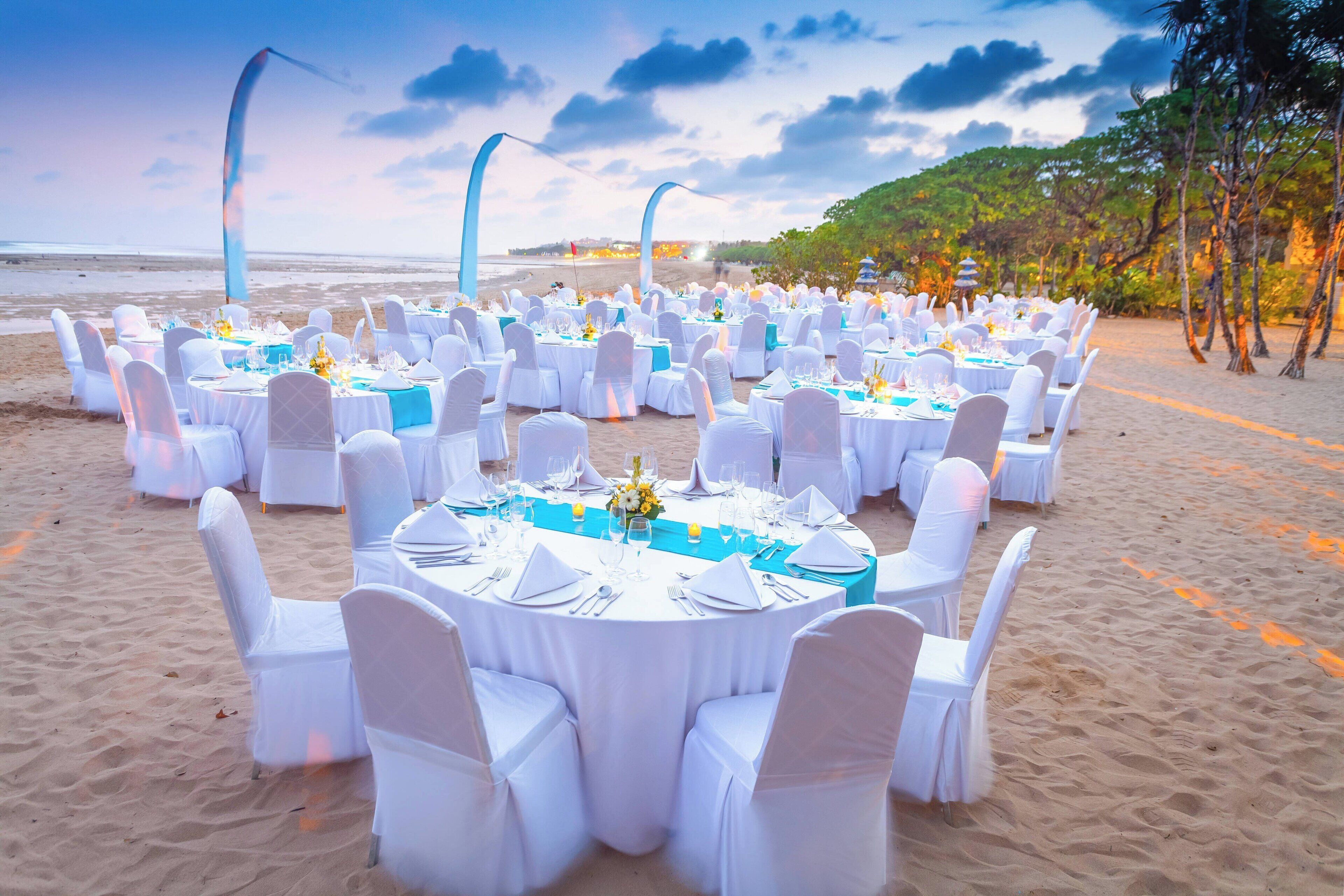 Montaje para una boda en la playa, con mesas redondas, sillas blancas y vista al océano.