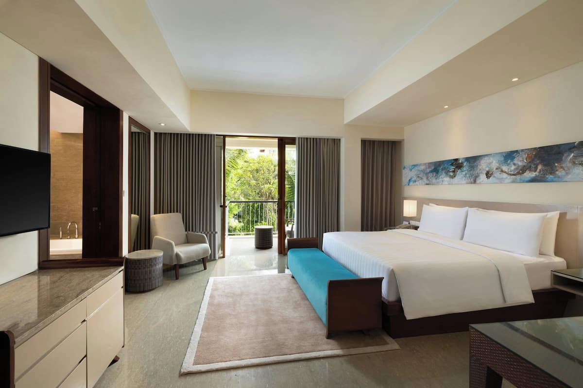 A hotel room with a bed, a TV, a sofa, a small table, and a view of greenery outside.