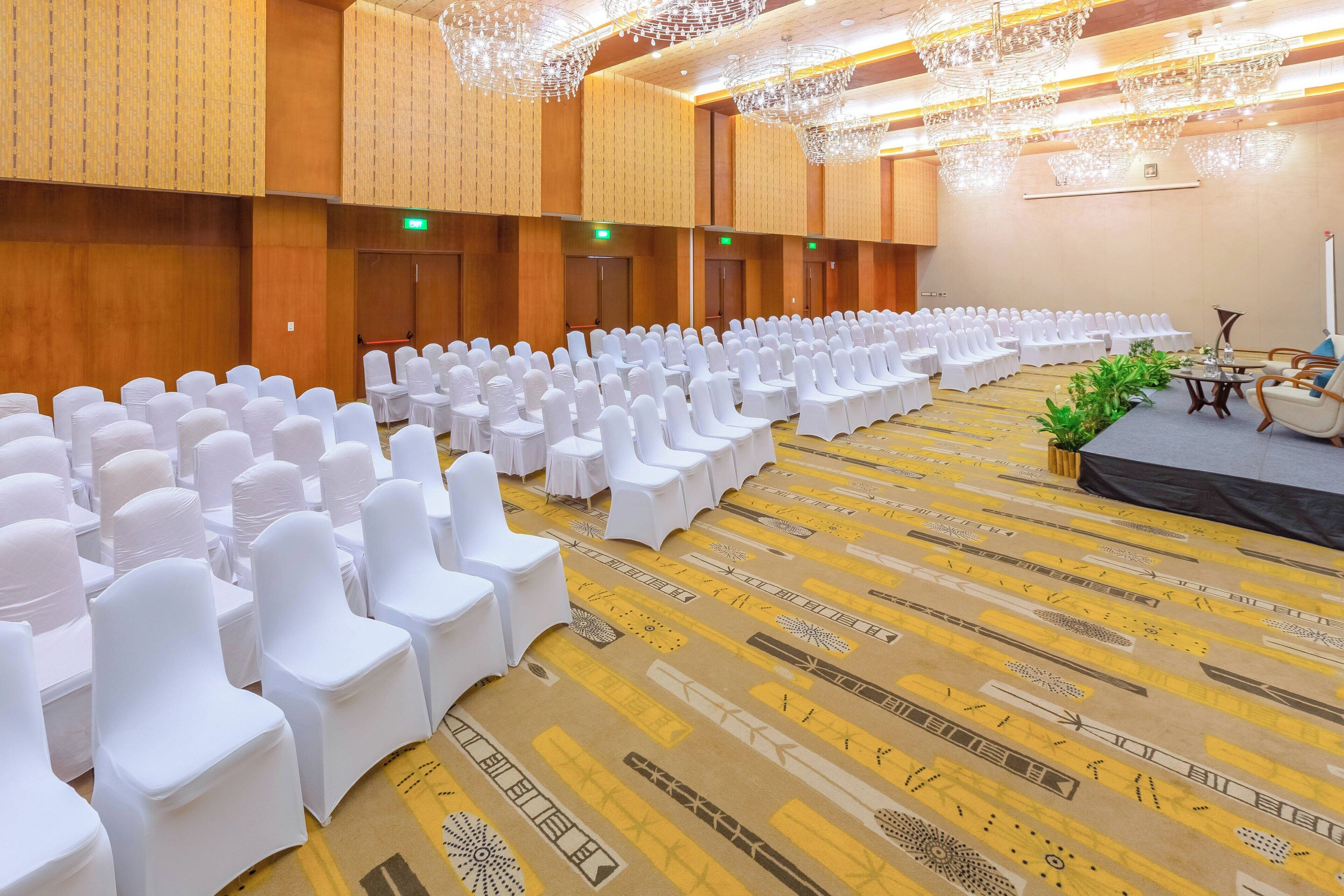 A large conference room with rows of white chairs and tables, wooden paneling on the walls, and a carpet with geometric patterns.