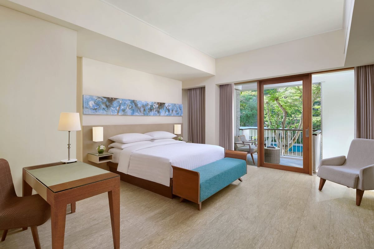 Quarto de hotel com cama, escrivaninha, cadeira, varanda e vista para o verde, além de um quadro na parede.