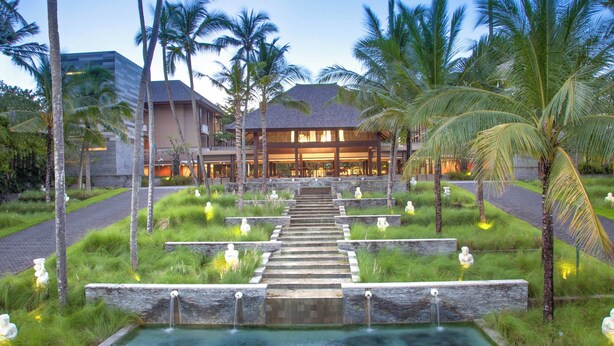 Een resort met een centraal waterpartij, palmbomen en een gebouw met schuine daken.