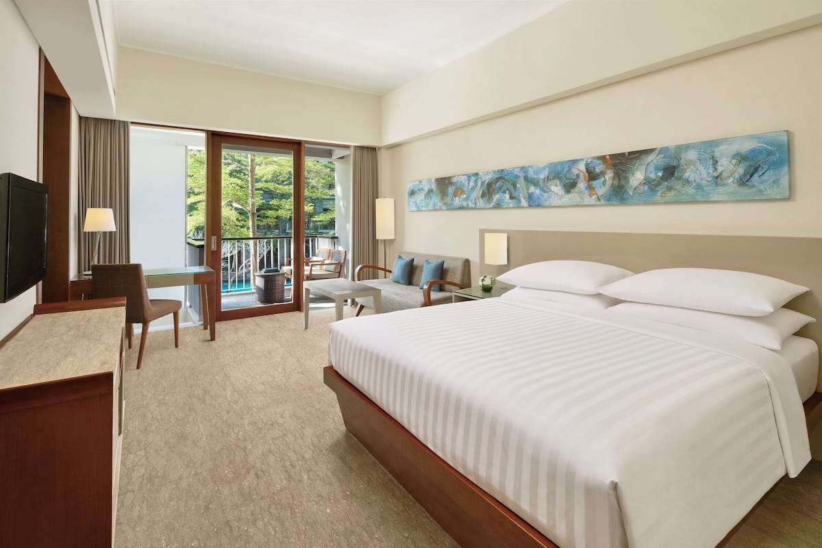 Quarto de hotel com uma cama grande, uma escrivaninha com cadeira, uma televisão e uma varanda com vista para árvores.