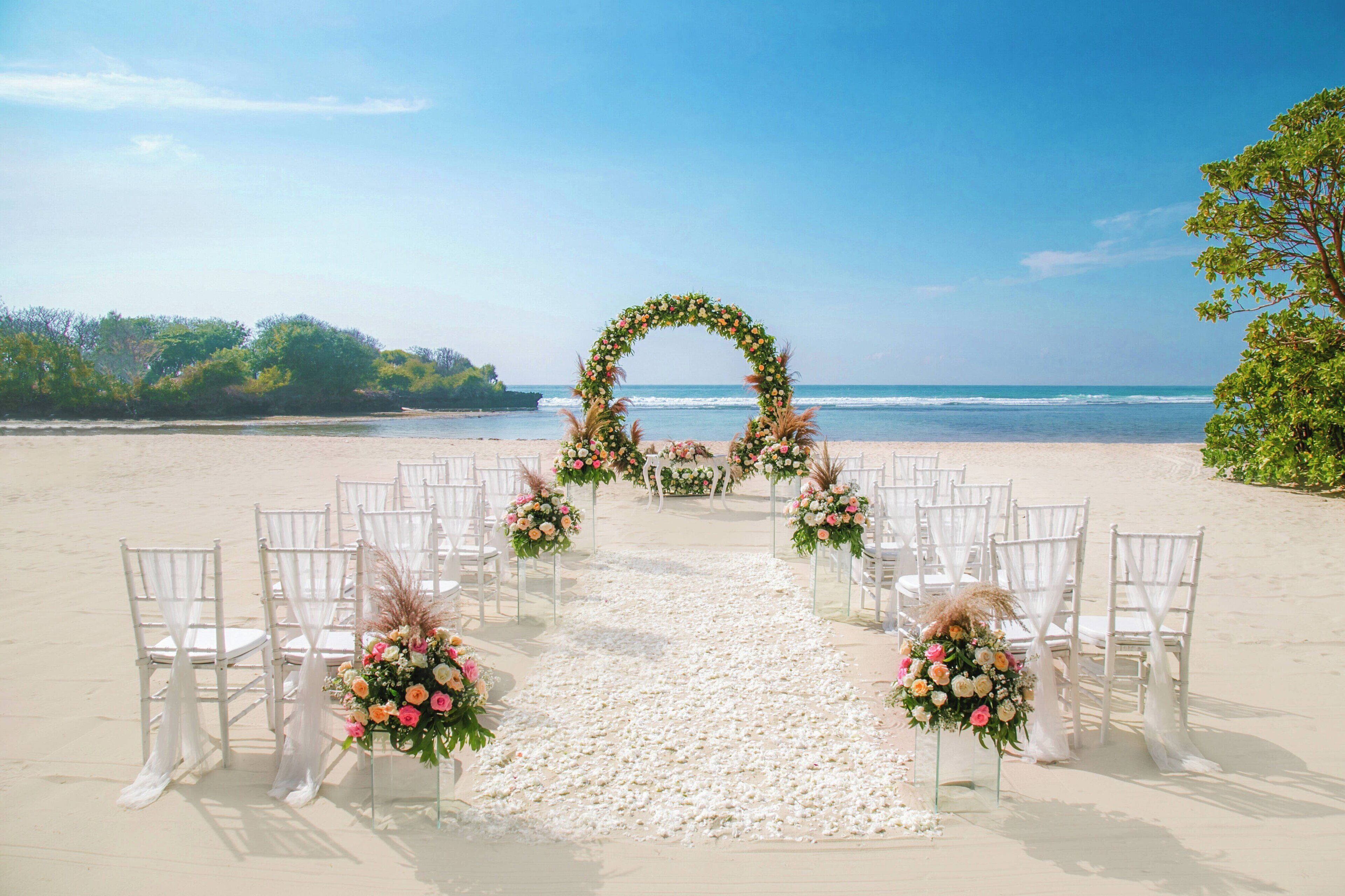 A beach wedding setup with white chairs and a floral arch.