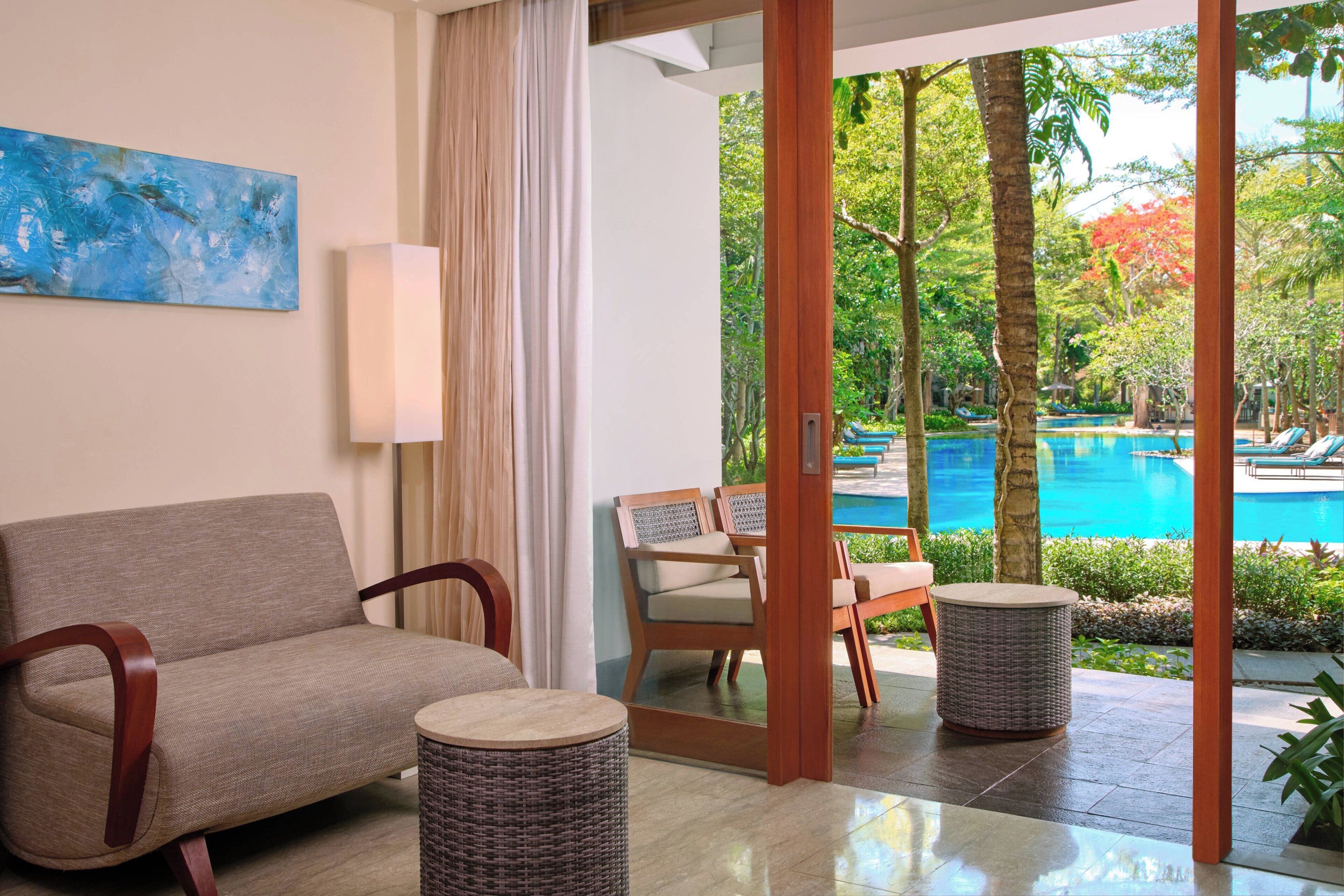 A room with a sofa, a chair, and a small table, with a view of a swimming pool and trees outside.