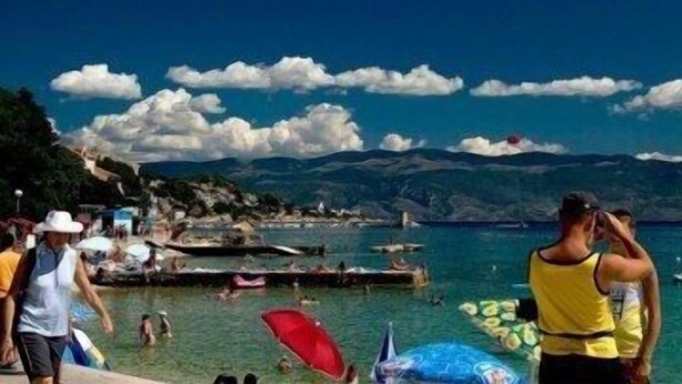 Una platja concorreguda amb gent, barques i un paisatge de muntanyes al fons.