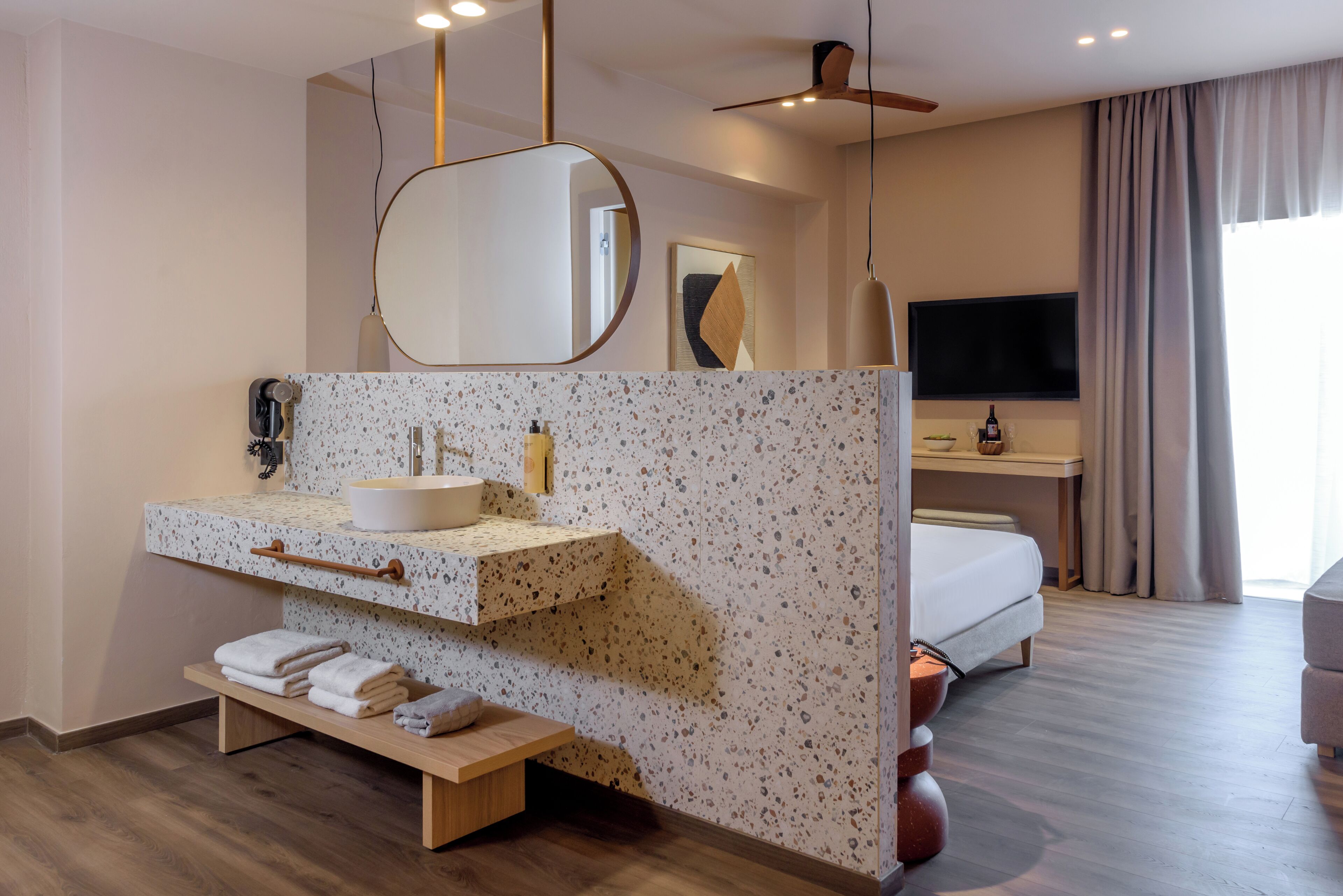 A modern hotel room with a large mirror, a sink, a wooden shelf with towels, a bed, a TV, and a ceiling fan.