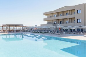 A pool area with lounge chairs and umbrellas, a covered seating area, and a multi-story building with balconies. - Zeus Hotels Neptuno Beach - All Inclusive (Malevizi)