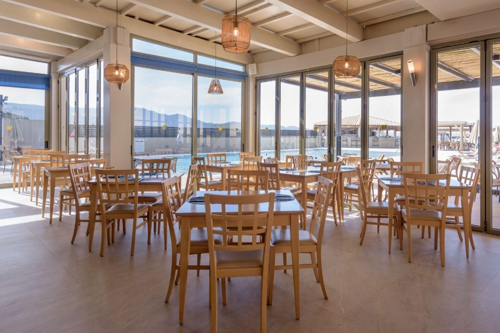 A spacious dining area with wooden tables and chairs, large windows offering a view of a pool and mountains.