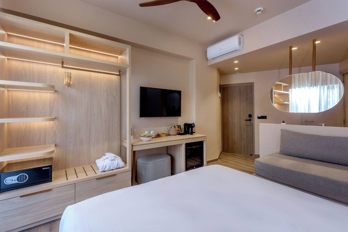 A hotel room with a bed, a desk with a flat-screen TV, a minibar, a seating area with a sofa, and built-in storage.