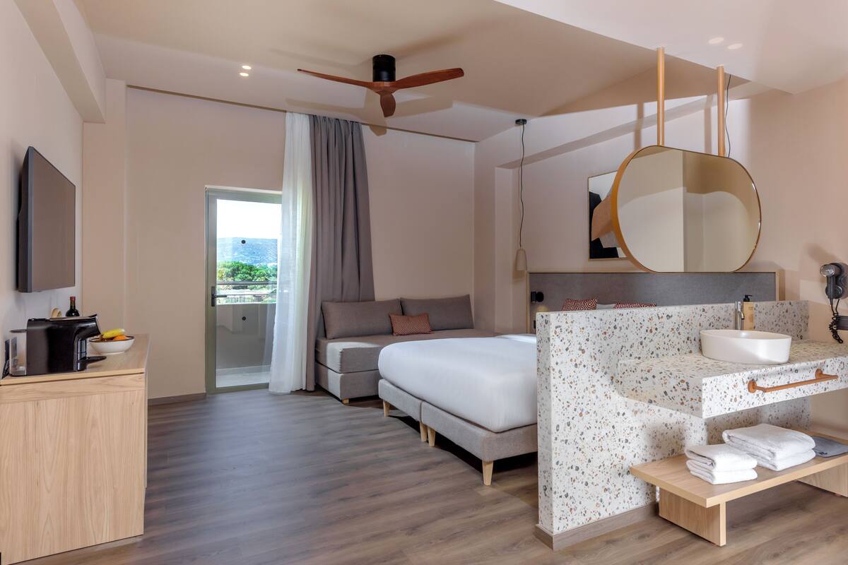 A modern hotel room with a bed, a desk, a bathroom with a sink and mirror, and a view of the outdoors.