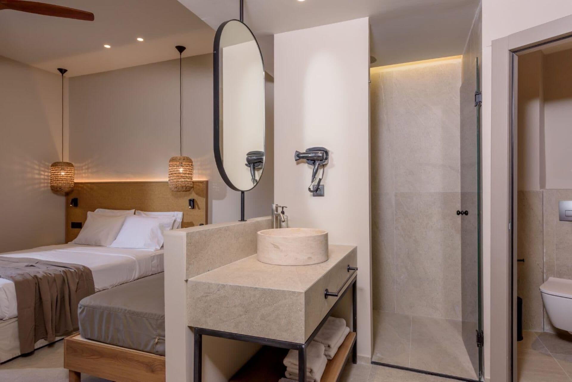 Una habitación de hotel moderna con una cama grande, una mesita de noche, un lavamanos redondo, un espejo, un secador de pelo y una ducha a ras de suelo.