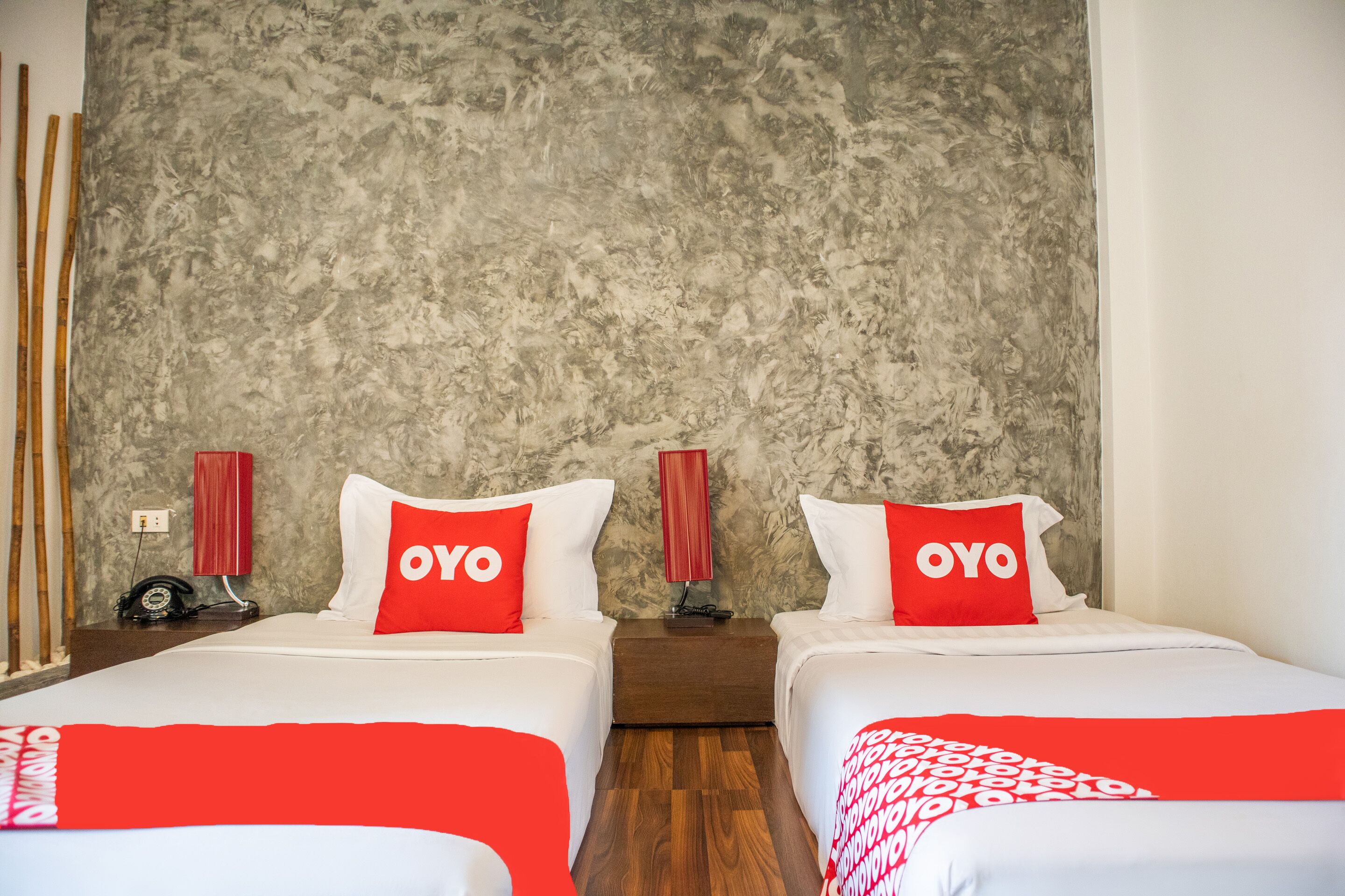 A hotel room with two beds, each with a red 'OYO' pillow, a red lamp on the nightstand, and a textured gray wall.