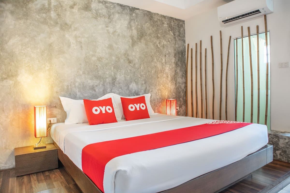 Un lit recouvert d’une couvre-lit rouge et blanche, deux oreillers portant le logo « OYO », une table de chevet avec une lampe, et un mur avec des lattes de bois verticales.