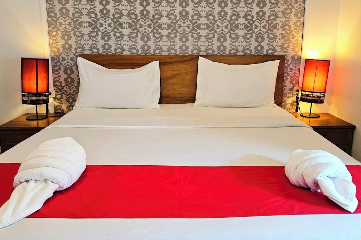 Un lit bien fait, avec du linge de lit blanc et deux oreillers, flanqué de deux lampes de chevet à abat-jour rouge, le tout mis en valeur par un papier peint à motifs.