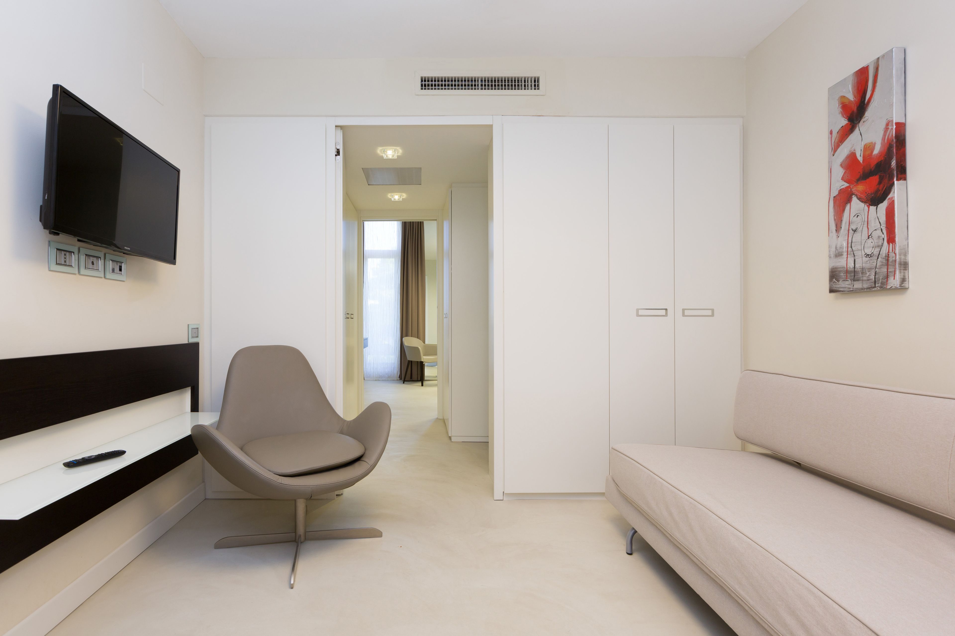 Une chambre d’hôtel moderne équipée d’un téléviseur à écran plat fixé au mur, d’un canapé confortable, d’un fauteuil élégant et d’une porte donnant sur un couloir.