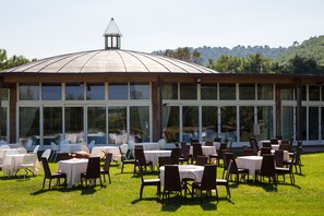 An outdoor dining area with round tables and chairs, a large glass building with a dome roof, and a view of a forested hillside. - Gusmay Resort – Hotel Gusmay & Suite Le Dune (Peschici)
