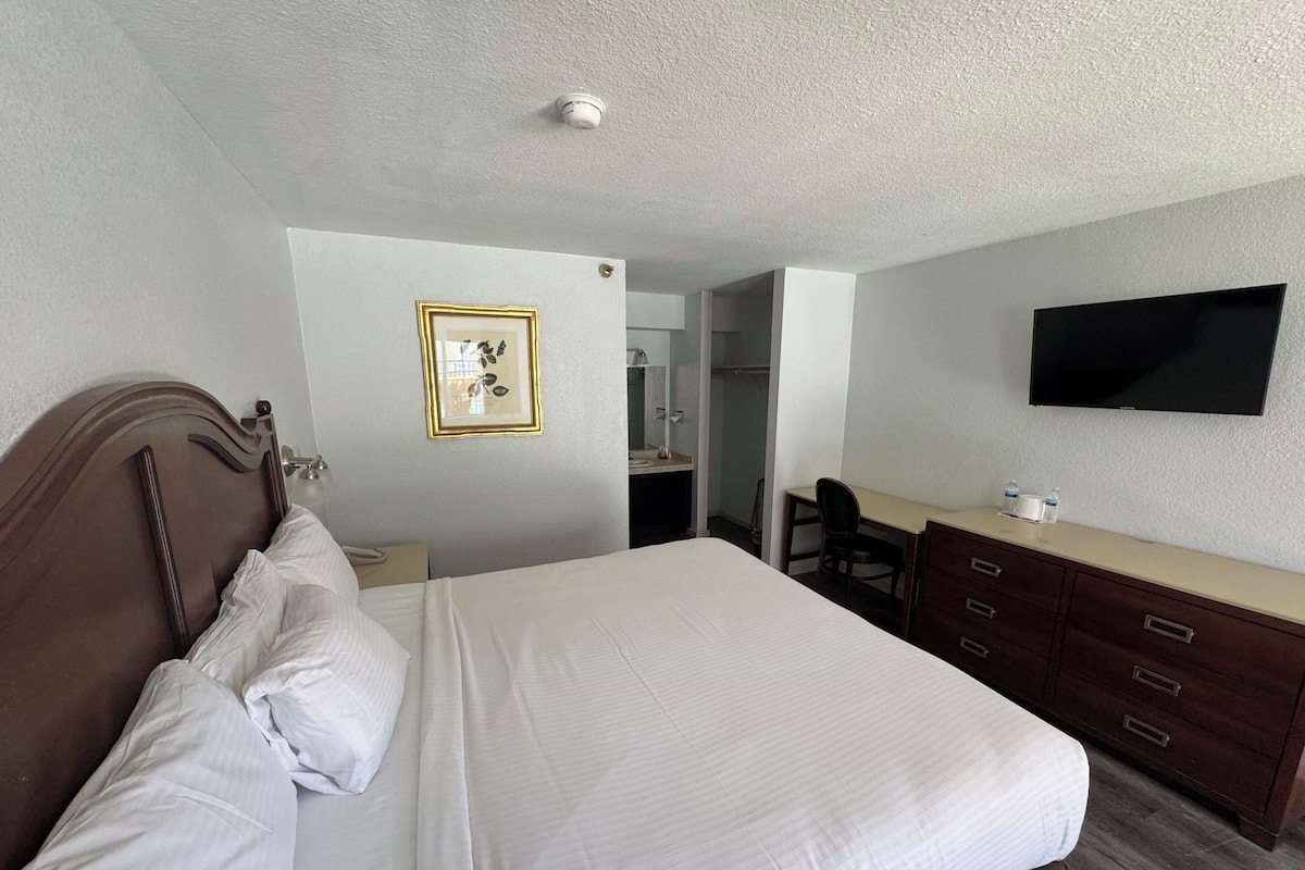 A hotel room with a bed, a desk, a chair, a dresser, a television, and a bathroom visible through an open door.
