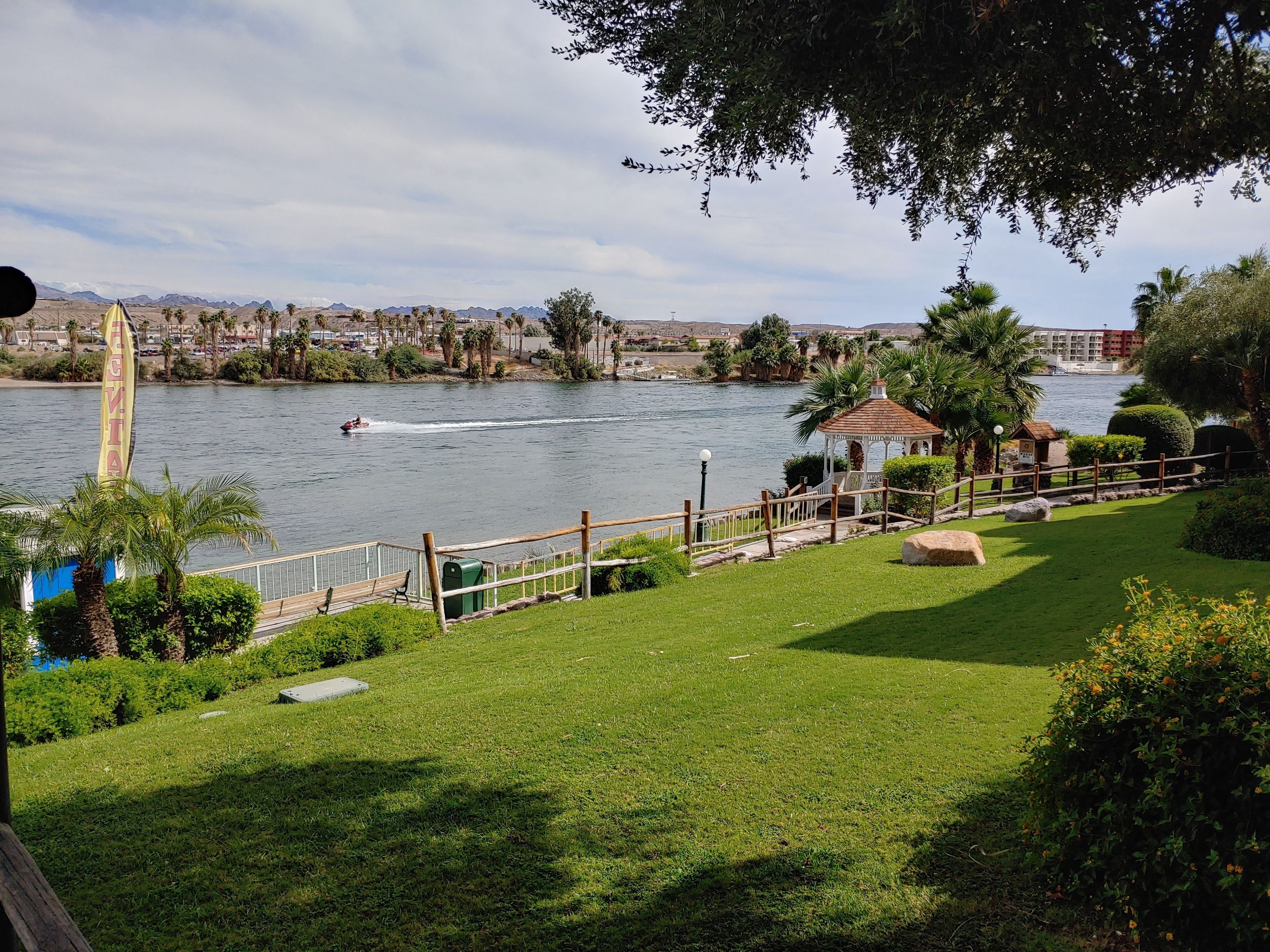 Vista para o rio com um coreto, um barco na água e uma área de parque com cerca e bancos.