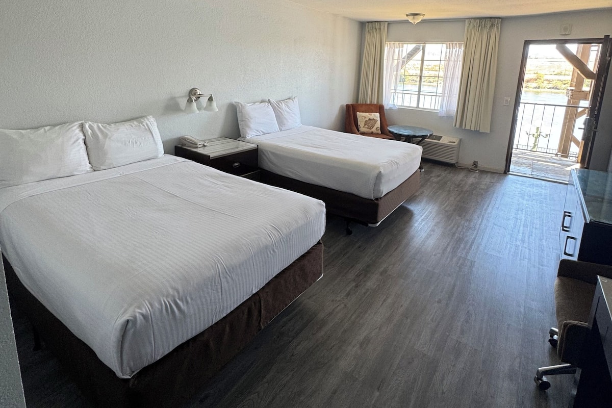 A hotel room with two beds, a desk, a chair, a window with curtains, and a view of the outdoors.