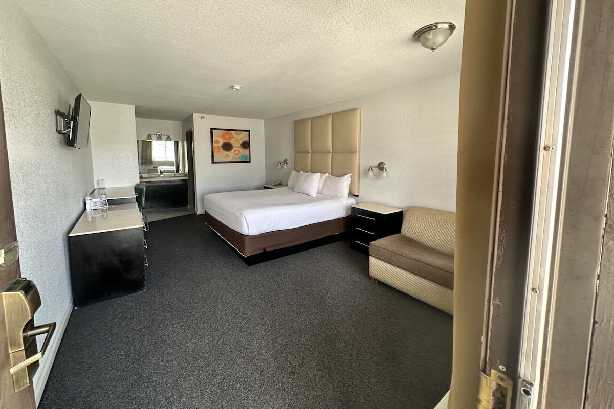 A hotel room with a bed, a desk, a chair, a small sofa, a nightstand, and a wall-mounted lamp.