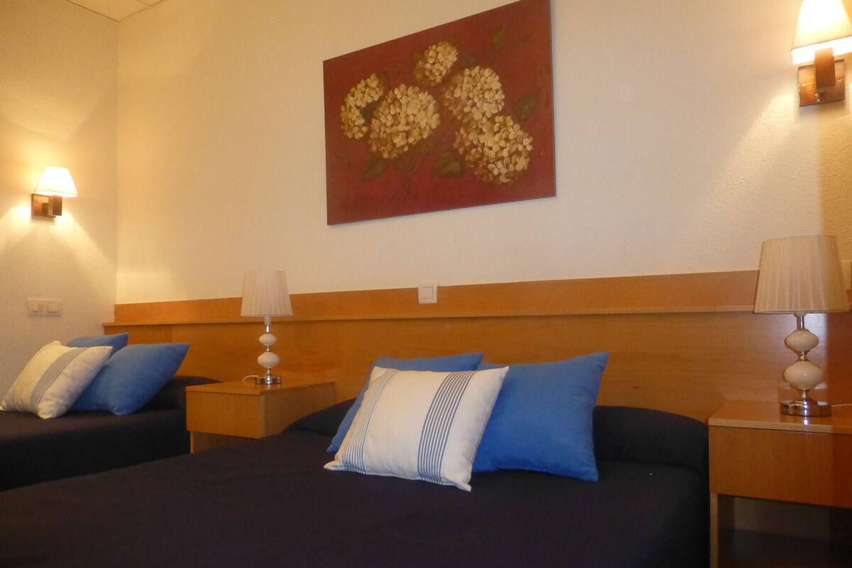 A hotel room with two beds, each with blue and white pillows, and a wooden headboard. There are two bedside lamps and a painting on the wall.