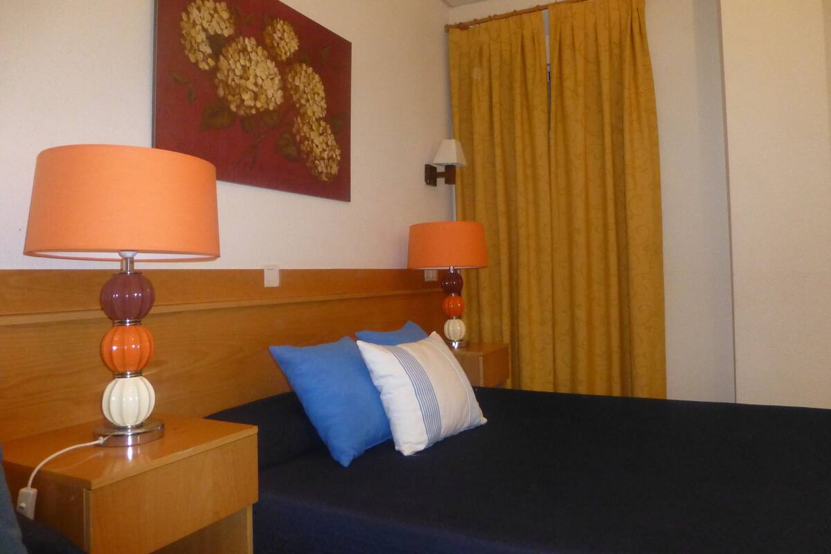A hotel room with a bed, two bedside lamps, a painting on the wall, and a window with curtains.