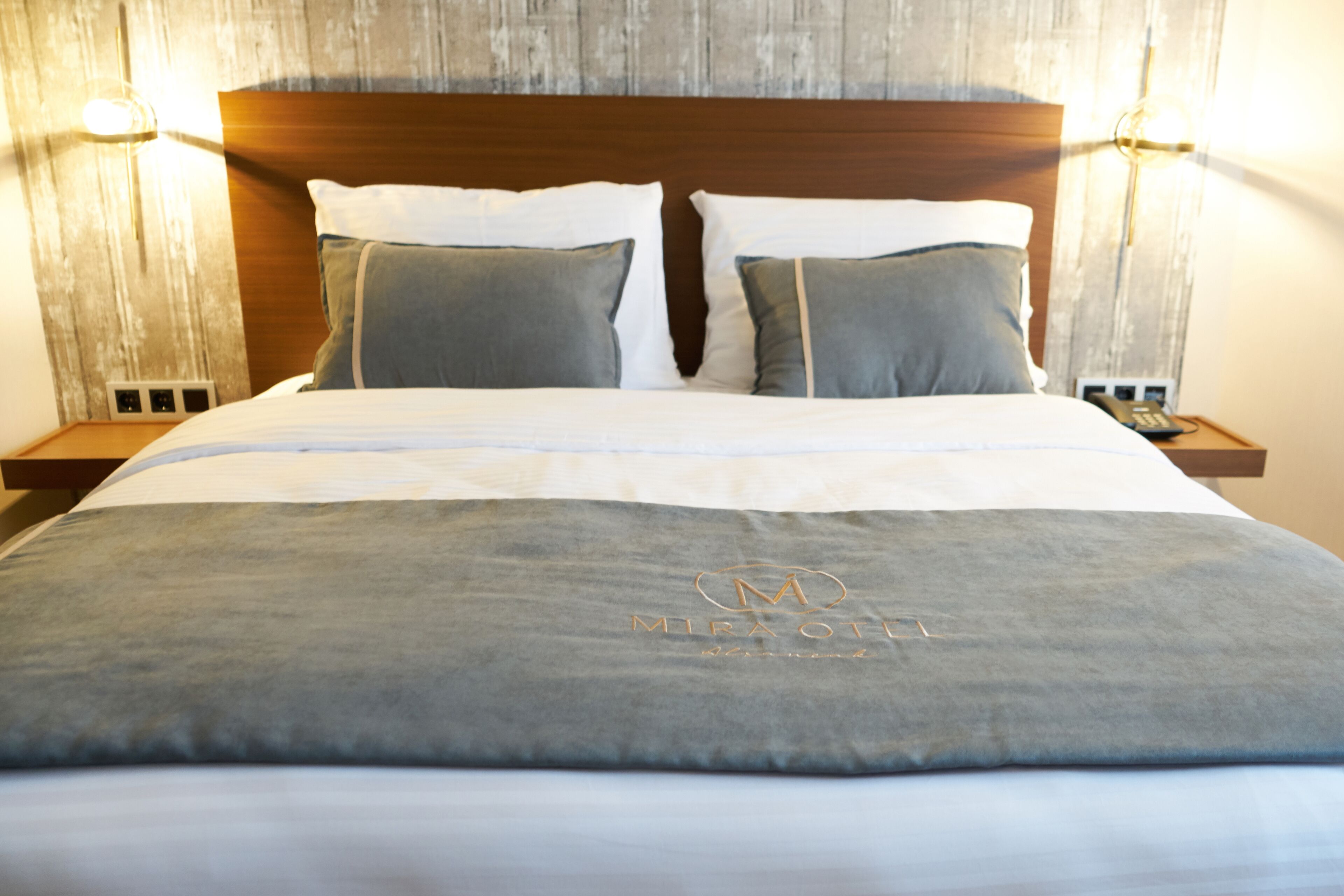Ein ordentlich bezogenes Bett mit grau-weißer Tagesdecke, zwei Kissen und einem Kopfteil mit Wandleuchten.