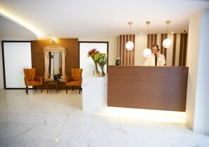 A hotel reception area with a marble floor, two orange chairs, a wooden reception desk, and a large mirror above the desk. - Mira Otel Alsancak (Izmir)