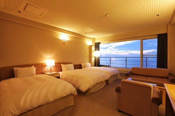 A hotel room with two beds, a sofa, a table, and a large window with a view of the sea.