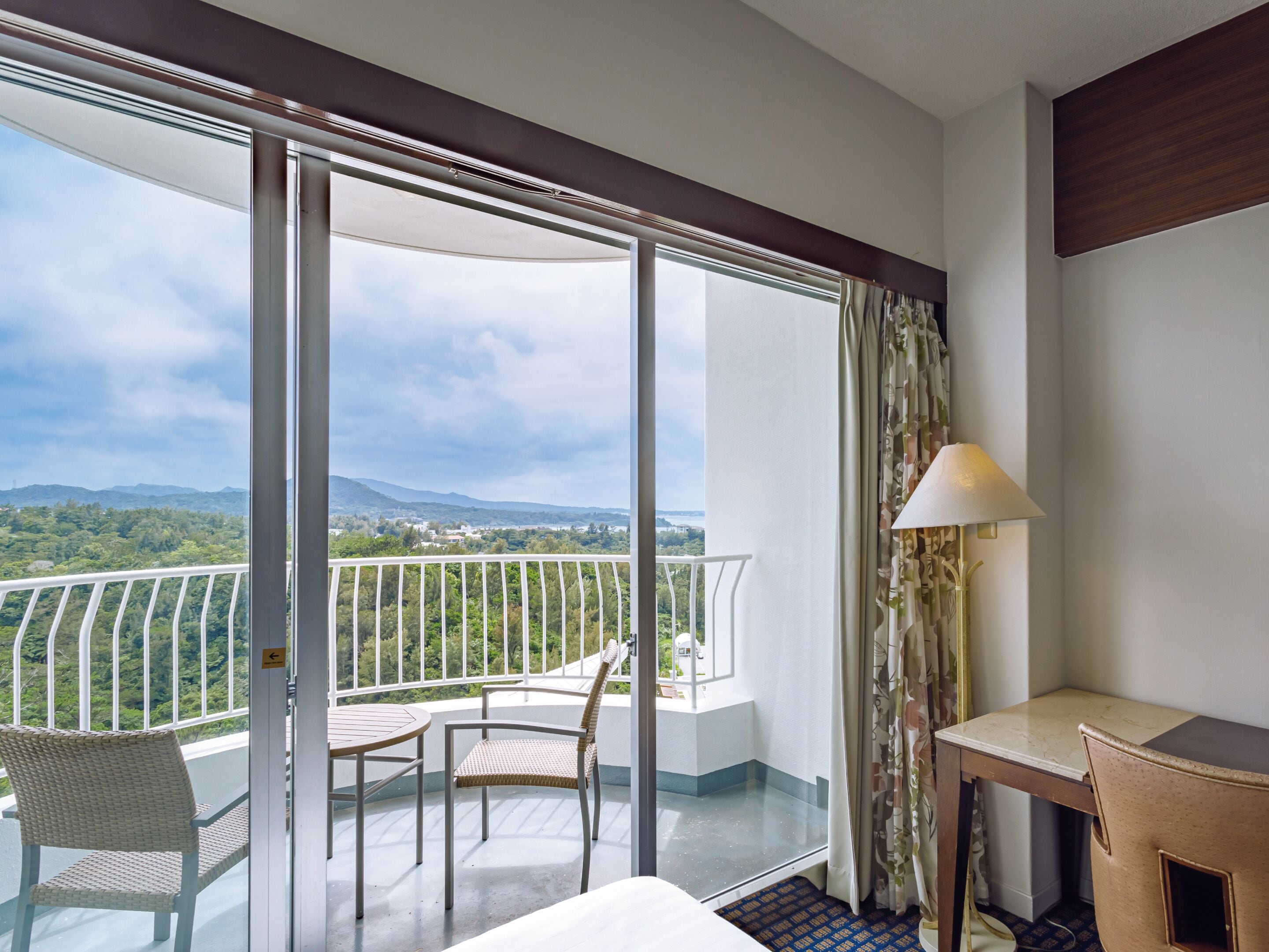 A hotel room with a balcony, a table, a chair, and a lamp.