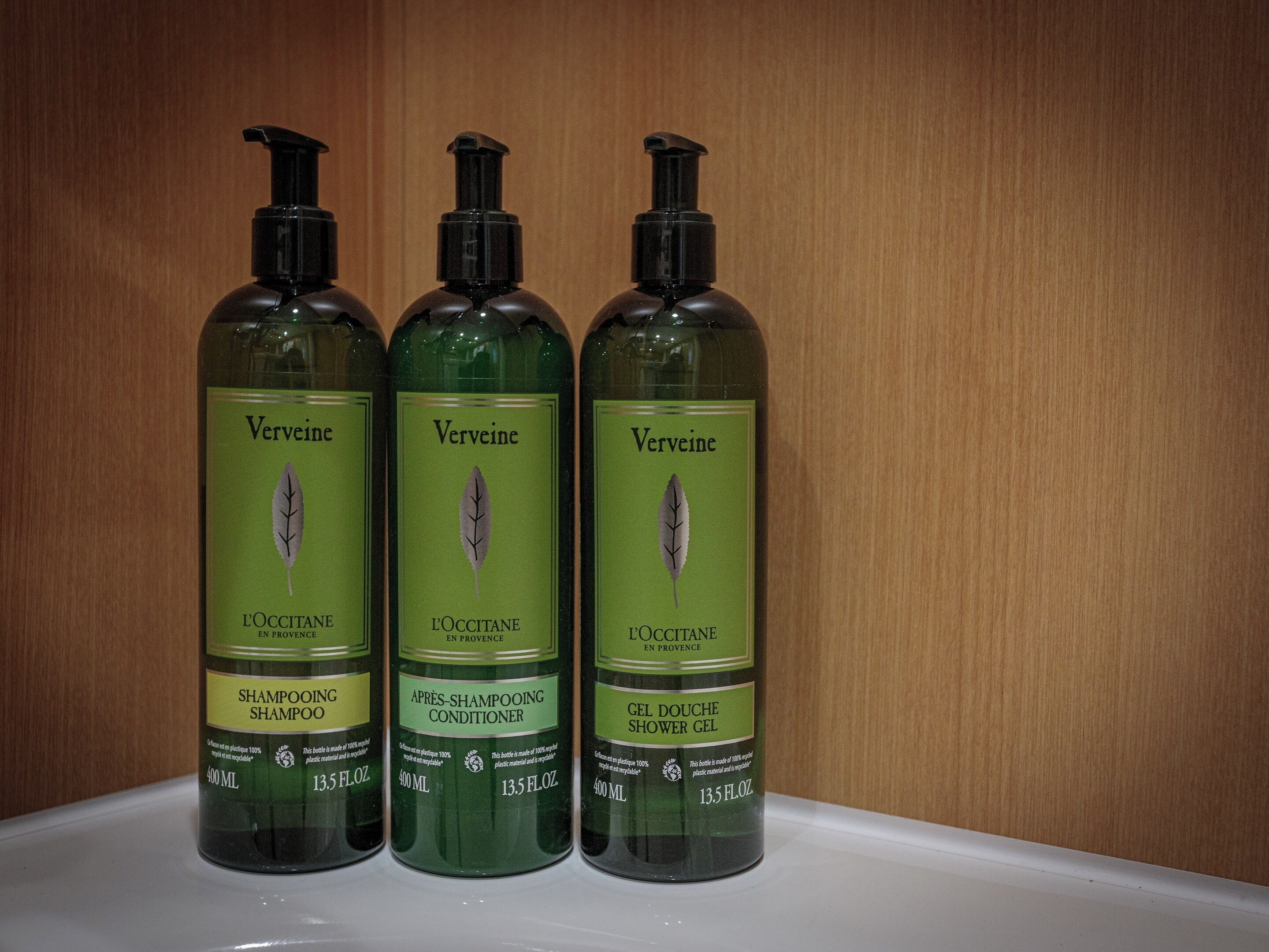 Drie flessen Verveine shampoo en conditioner op een wit oppervlak, met een houten achtergrond.