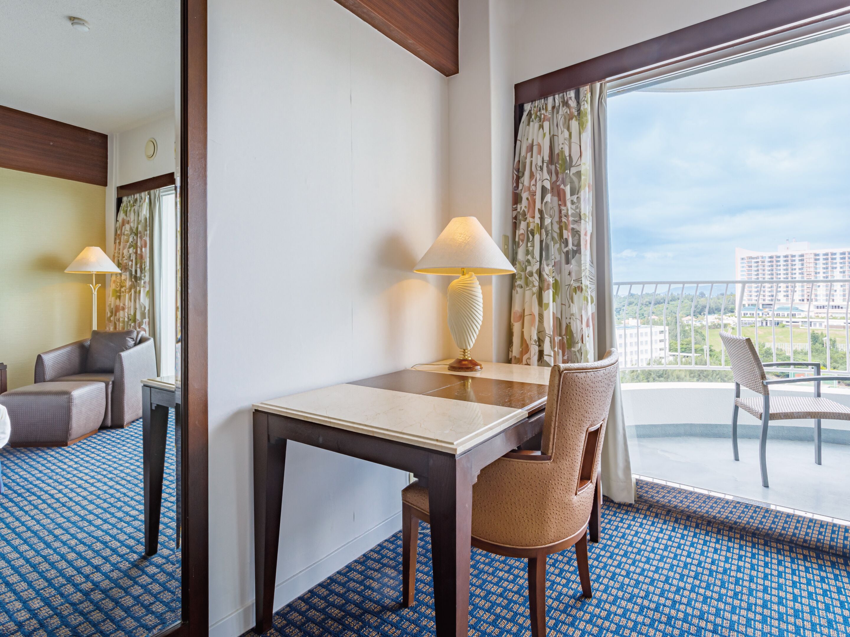 A hotel room with a balcony, a desk, two chairs, and a lamp.