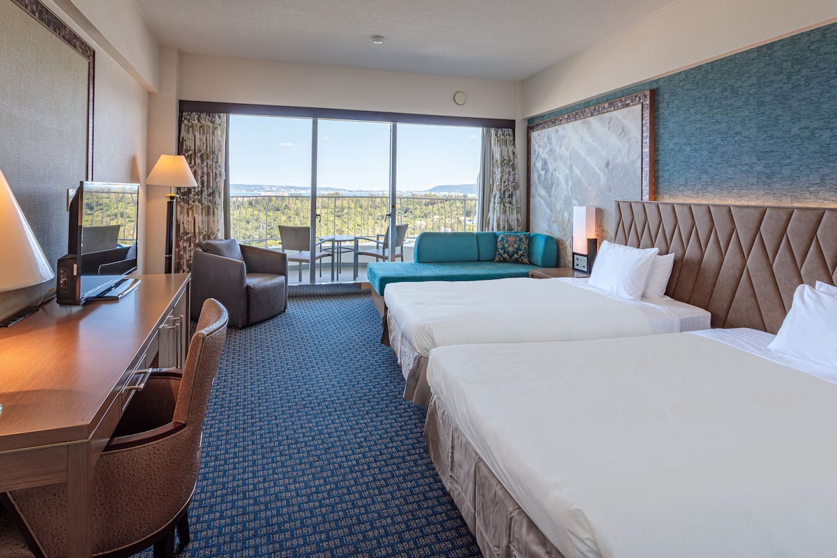 Habitación de hotel con dos camas, un escritorio, televisión y balcón con vistas.